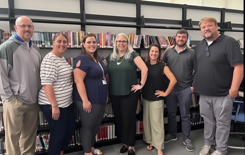 A diverse group of seven people, staff of ms blanchard joseph a foran, stand smiling in front of a wall of tall, black metal bookshelves filled with various books. They are all casually dressed